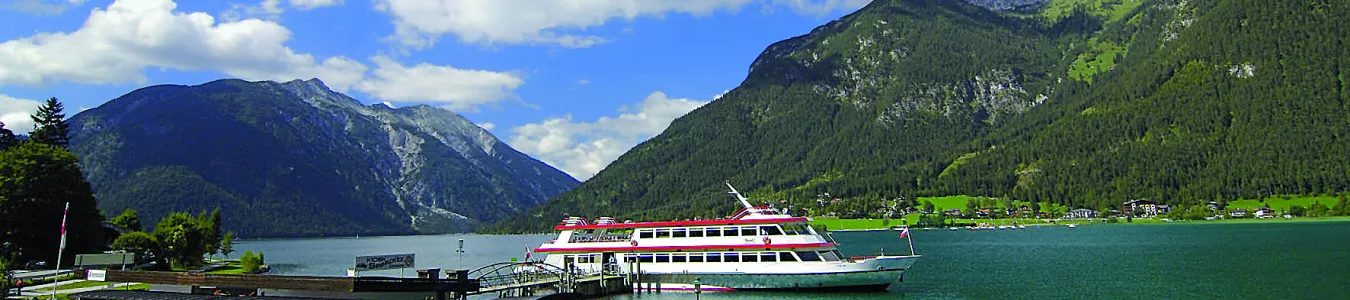 Blick auf einen See, umgeben von Bergen, mit Booten und Menschen am Ufer.
