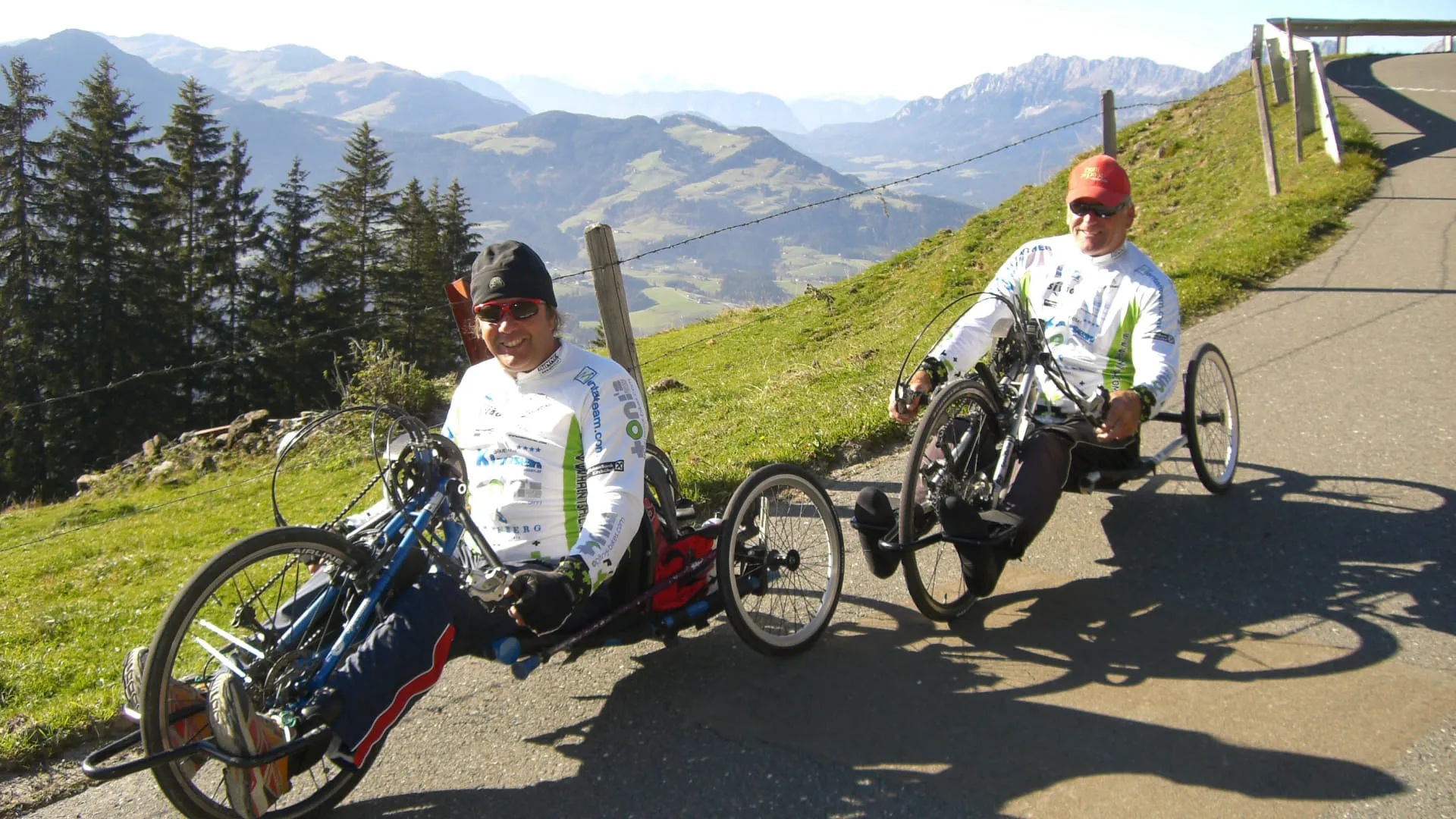 Zwei Männer auf Handbikes fahren einen Bergweg mit bergiger Landschaft im Hintergrund.